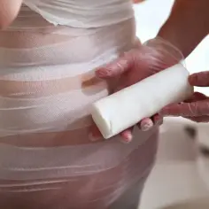 Osoba w rękawiczkach trzyma biały wałek, podczas gdy druga osoba owija ciało bandażem. Widoczne body wrapping efekty.
