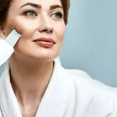 Kobieta w białym szlafroku wykonuje zabieg ultradźwiękami. Widoczne są **peeling kawitacyjny efekty** – gładka, promienna skóra.