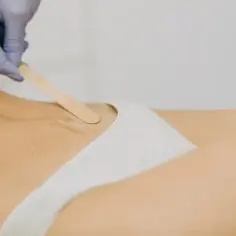 Ręka w rękawiczce aplikuje wosk do depilacji odbytu drewnianą szpatułką.