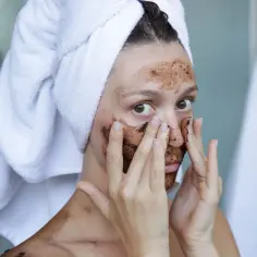 Kobieta z ręcznikiem na głowie aplikuje na twarz domowy peeling do twarzy z kawy.