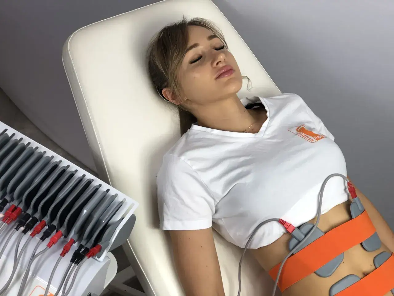 Kobieta leży na kozetce, poddana elektrostymulacji brzucha. Urządzenie z elektrodami jest podłączone do jej ciała.